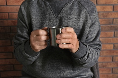 Photo of Poor elderly man holding mug near brick wall, focus on hands