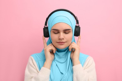 Muslim woman in hijab and headphones listening to music on pink background