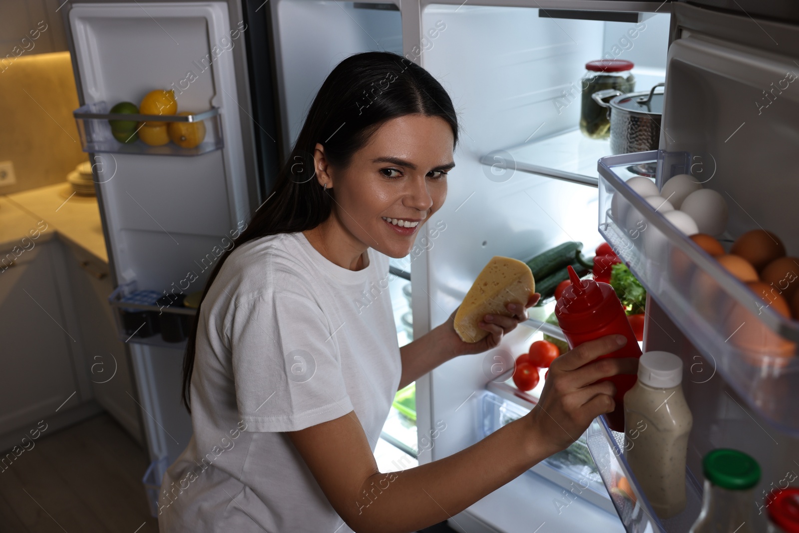 Photo of Young woman taking ketchup and cheese out of refrigerator in kitchen at night