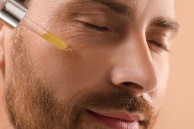 Handsome man applying cosmetic serum onto his face on beige background, closeup