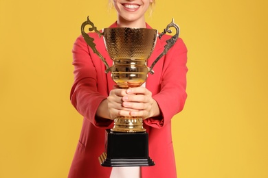 Photo of Young businesswoman with gold trophy cup on yellow background, closeup