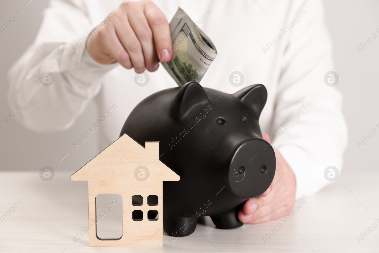 Photo of Man putting dollar banknotes into piggy bank near house model at wooden table, closeup. Saving money concept