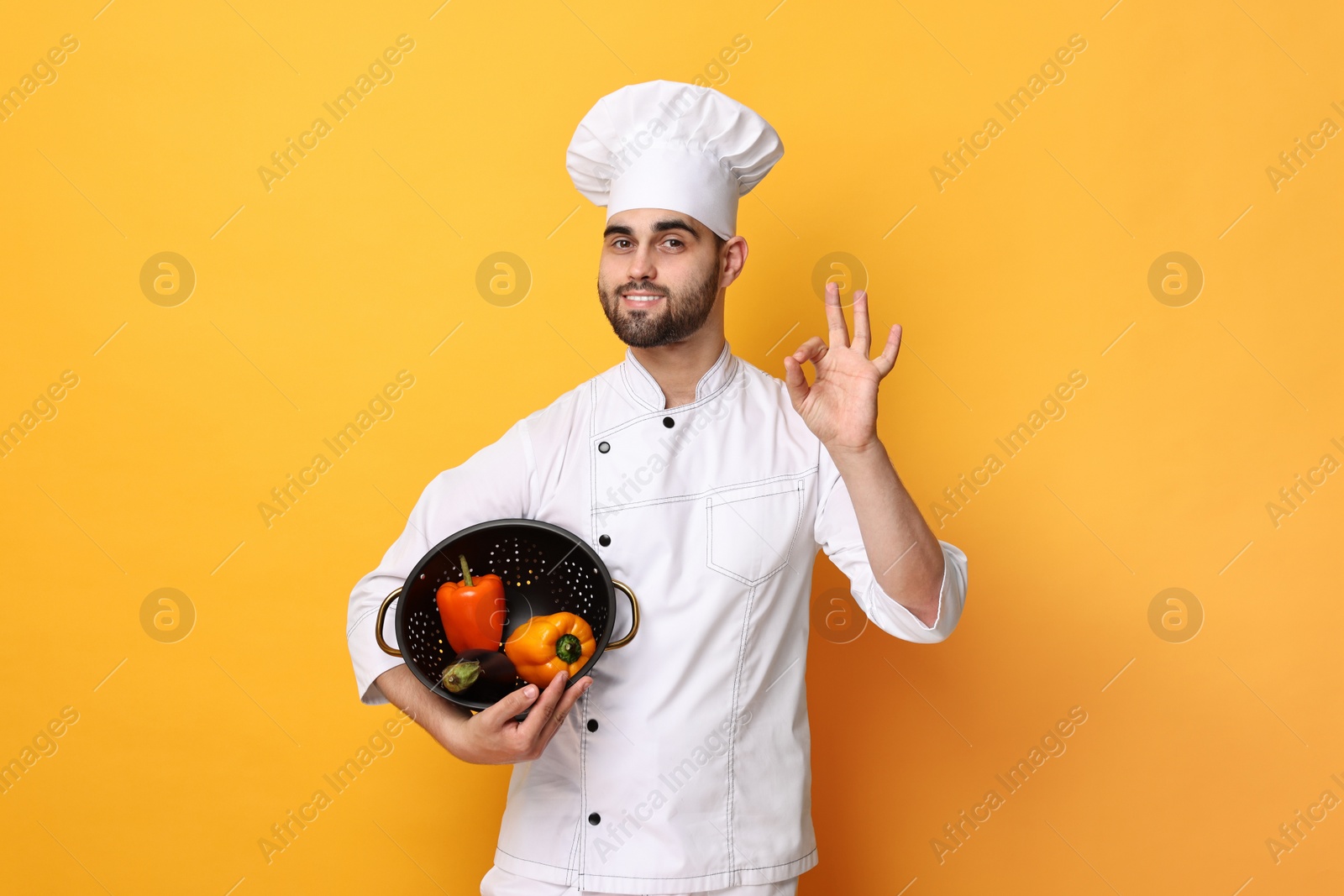 Photo of Professional chef holding colander with vegetables and showing OK gesture on yellow background
