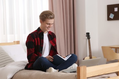 Online learning. Smiling teenage boy reading book near laptop on bed at home