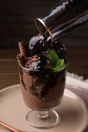 Photo of Adding chocolate topping into delicious ice cream with wafer sticks, donut and mint in glass dessert bowl on table, closeup