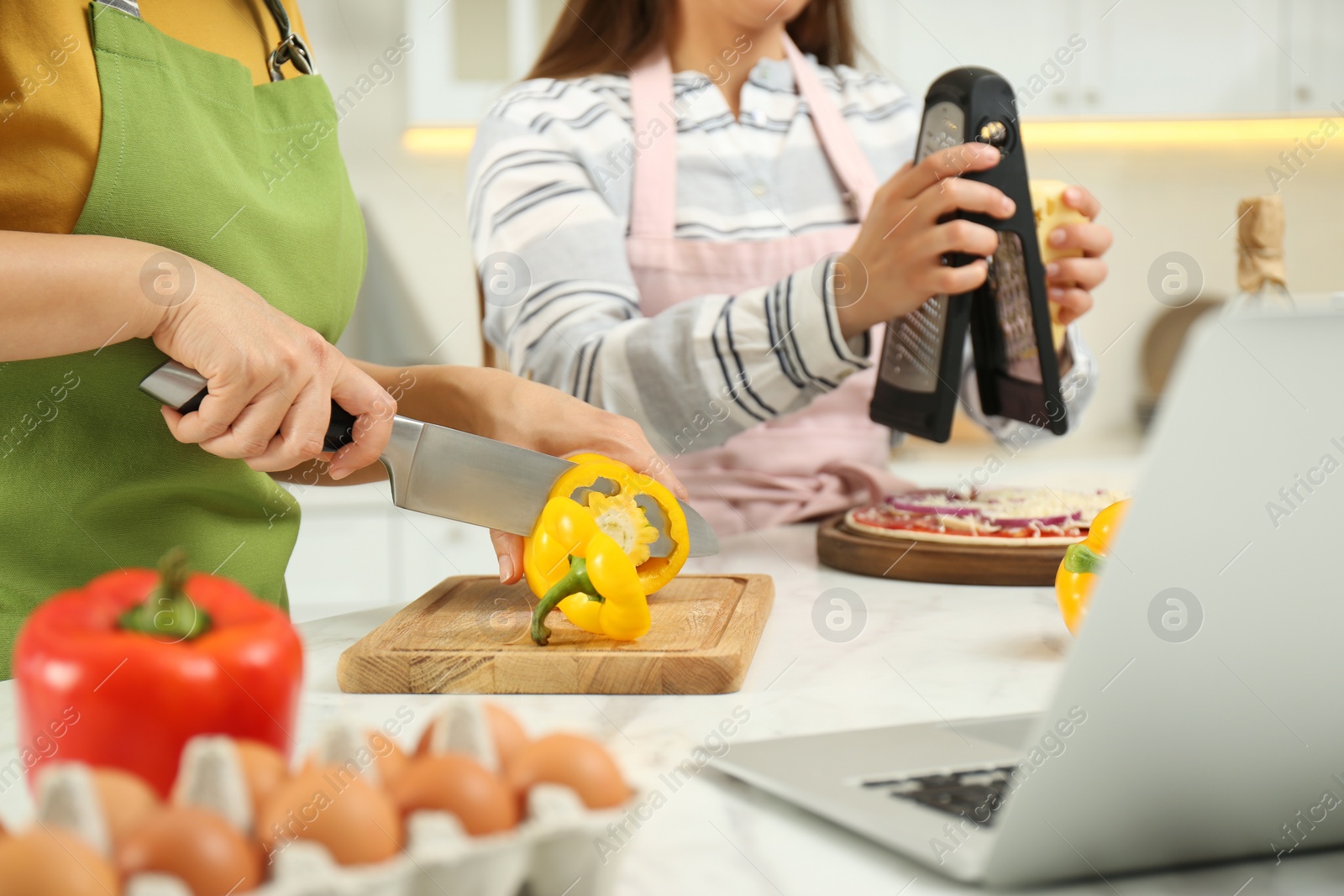 Photo of Mother with adult daughter making dinner while watching online cooking course via laptop in kitchen, closeup