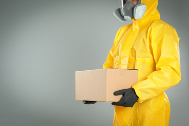 Photo of Man in chemical protective suit holding cardboard box on light grey background, closeup view with space for text. Prevention of virus spread
