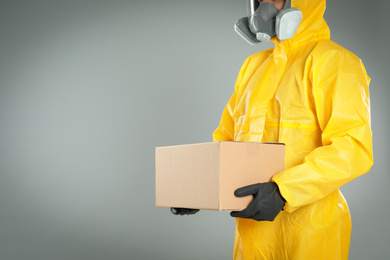 Man in chemical protective suit holding cardboard box on light grey background, closeup view with space for text. Prevention of virus spread