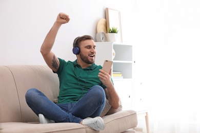 Photo of Young man in headphones with mobile device enjoying music on sofa at home