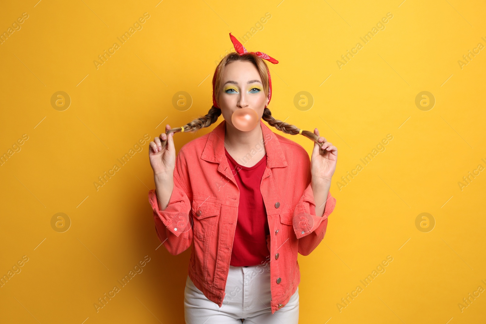 Photo of Fashionable young woman with braids and bright makeup blowing bubblegum on yellow background