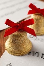 Photo of Golden shiny bells with red bows and music sheet on wooden table, closeup. Christmas decoration