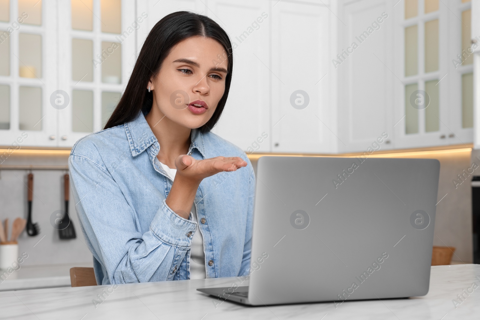 Photo of Happy young woman having video chat via laptop and blowing kiss at table in kitchen