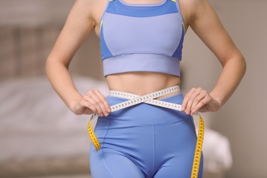 Photo of Woman in sportswear measuring waist with tape at home, closeup