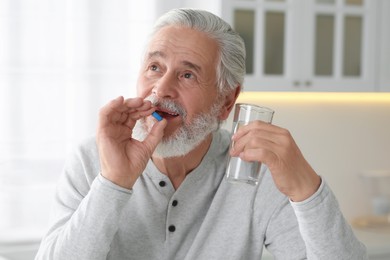 Photo of Senior man with glass of water taking pill at table in kitchen
