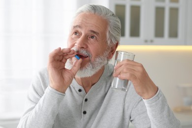 Senior man with glass of water taking pill at table in kitchen