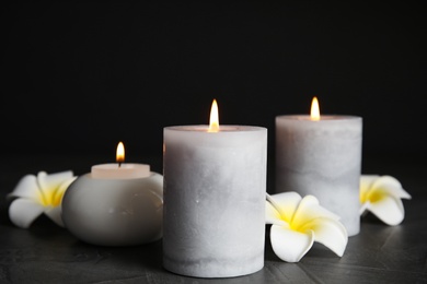 Photo of Burning candles and plumeria flowers on dark grey table against black background