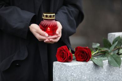 Woman holding candle near light grey tombstone with red roses outdoors, closeup. Funeral ceremony