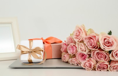 Photo of Beautiful bouquet of roses, gifts and laptop on white table. Happy birthday greetings