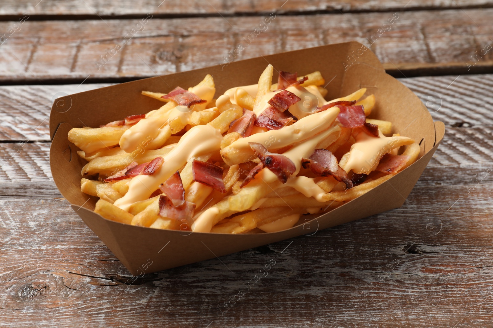 Photo of Tasty potato fries, cheese sauce and bacon in paper container on wooden table, closeup