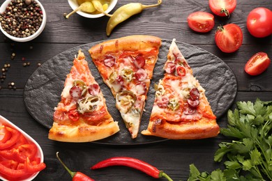 Photo of Slate board with pieces of delicious pizza Diablo among products on black wooden table, flat lay