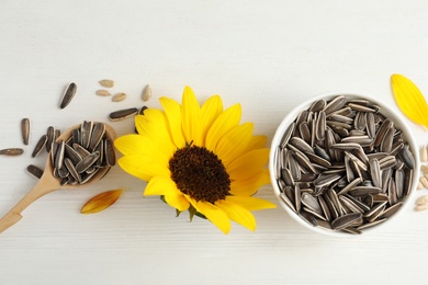 Photo of Organic sunflower seeds, spoon and bright flower on white wooden table, flat lay