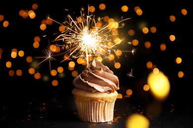 Delicious birthday cupcake with sparkler on black table against blurred lights