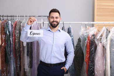 Photo of Dry-cleaning service. Happy worker holding Open sign near racks with clothes indoors