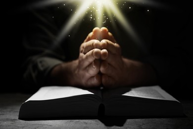 Image of Christian man praying over Bible in darkness, closeup. Holy light shining at him