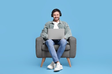 Photo of Happy man with laptop and headphones sitting in armchair on light blue background