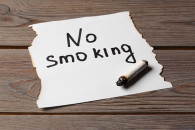 Photo of Burnt cigarette and words No Smoking written on paper on wooden table, closeup