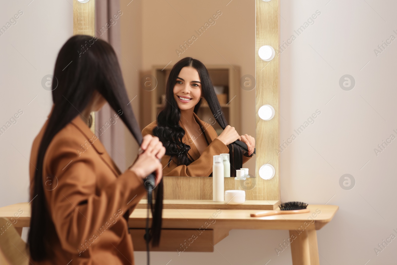Photo of Beautiful happy woman using hair iron near mirror in room