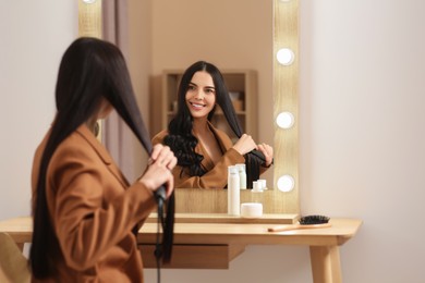 Beautiful happy woman using hair iron near mirror in room