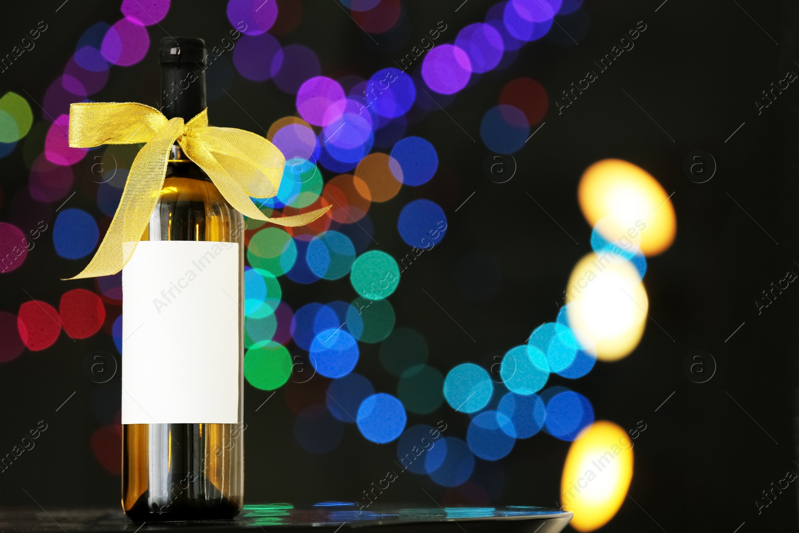 Photo of Bottle of wine with bow-knot on table against blurred lights, space for text. Bokeh effect