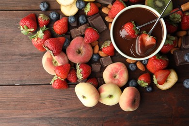 Fondue forks with strawberries in bowl of melted chocolate surrounded by other fruits on wooden table, flat lay