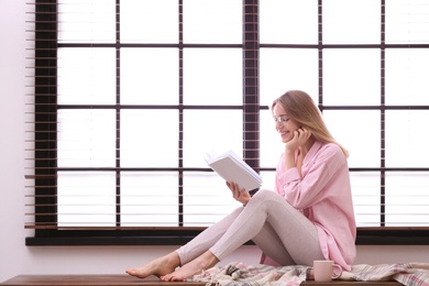 Young woman reading book near window with blinds at home. Space for text