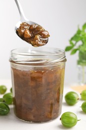 Photo of Spoon over jar with delicious gooseberry jam and fresh berries on white table, closeup