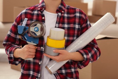 Photo of Woman with handheld dispenser, rolls of adhesive tape and plastic stretch wrap indoors, closeup