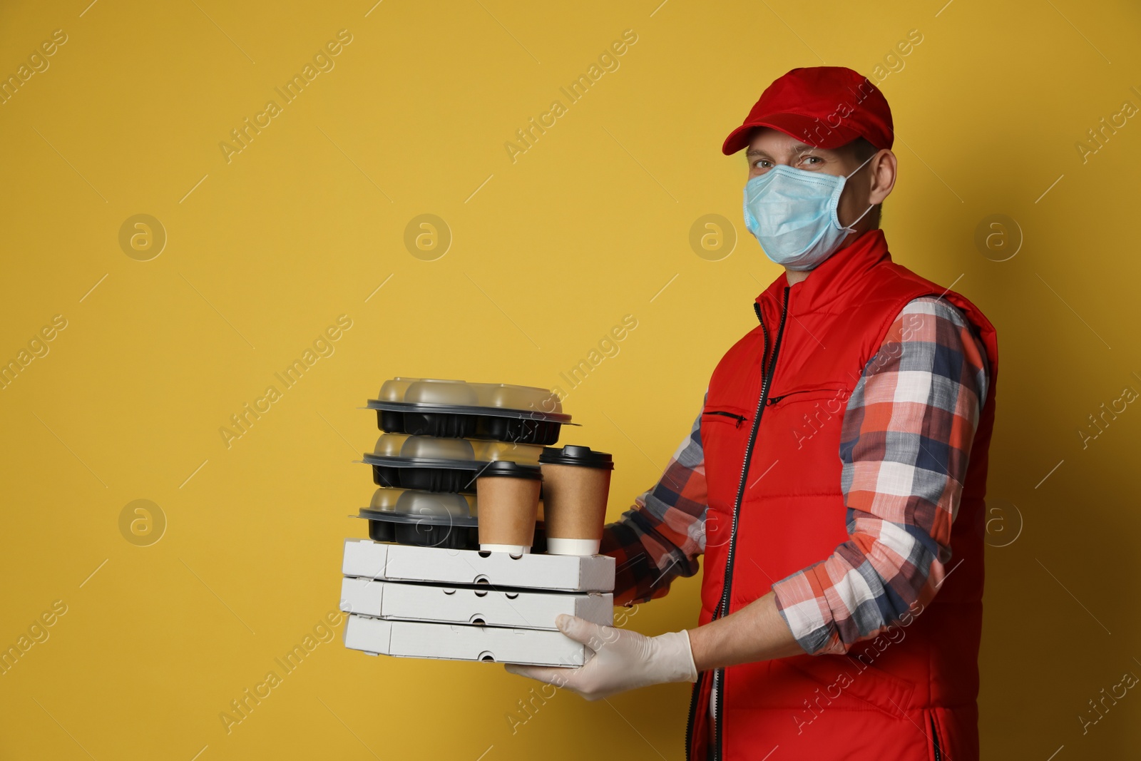 Photo of Courier in protective mask and gloves holding order on yellow background. Food delivery service during coronavirus quarantine