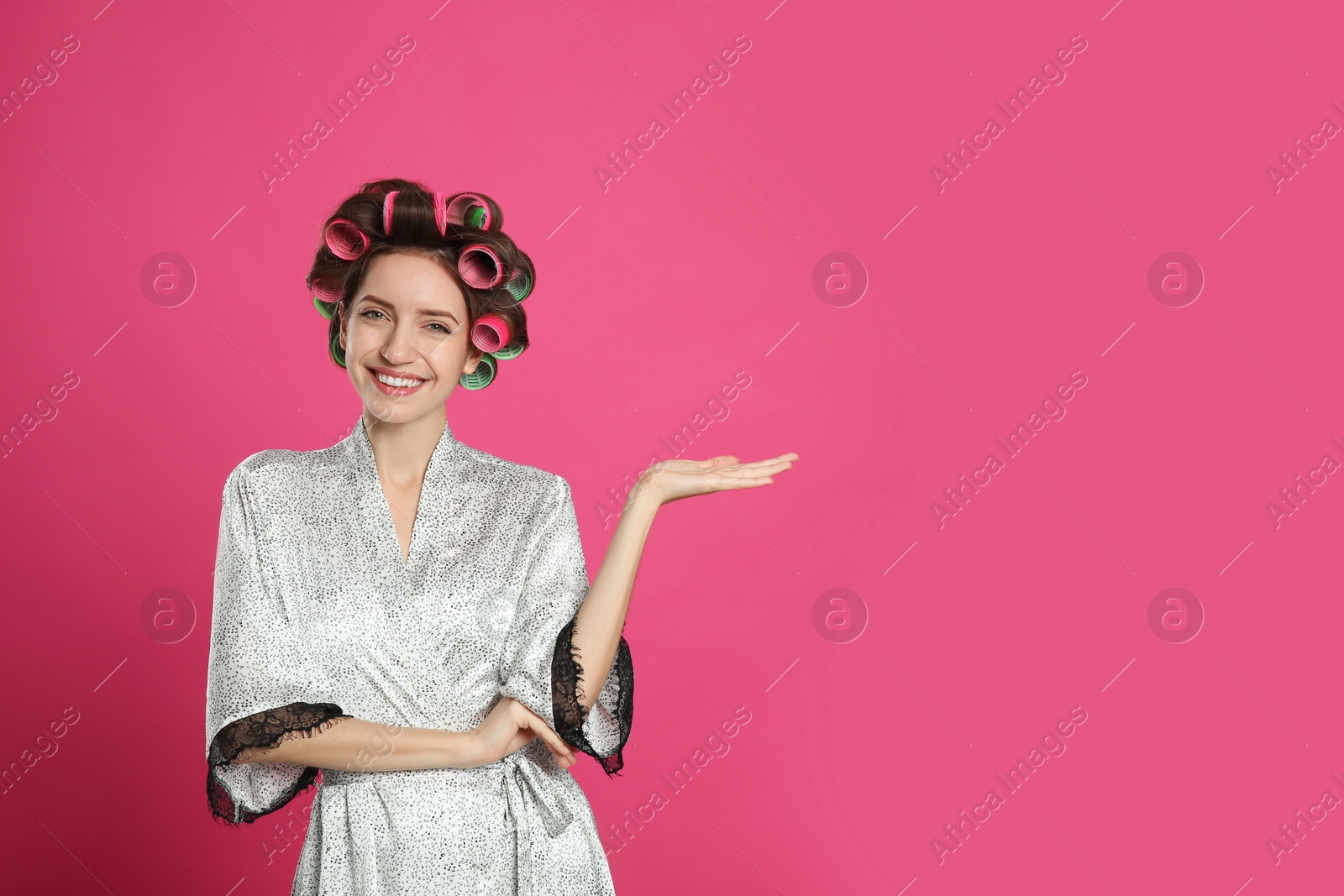 Photo of Beautiful young woman in silk bathrobe with hair curlers on pink background. Space for text