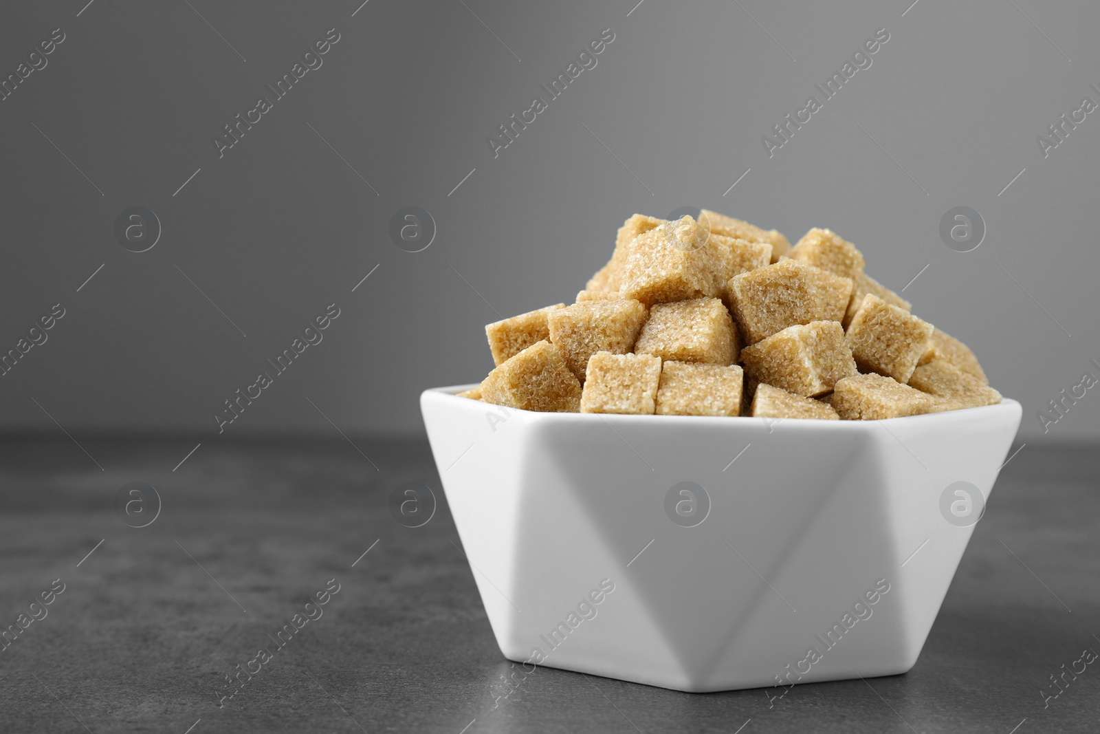 Photo of Brown sugar cubes in bowl on grey table. Space for text