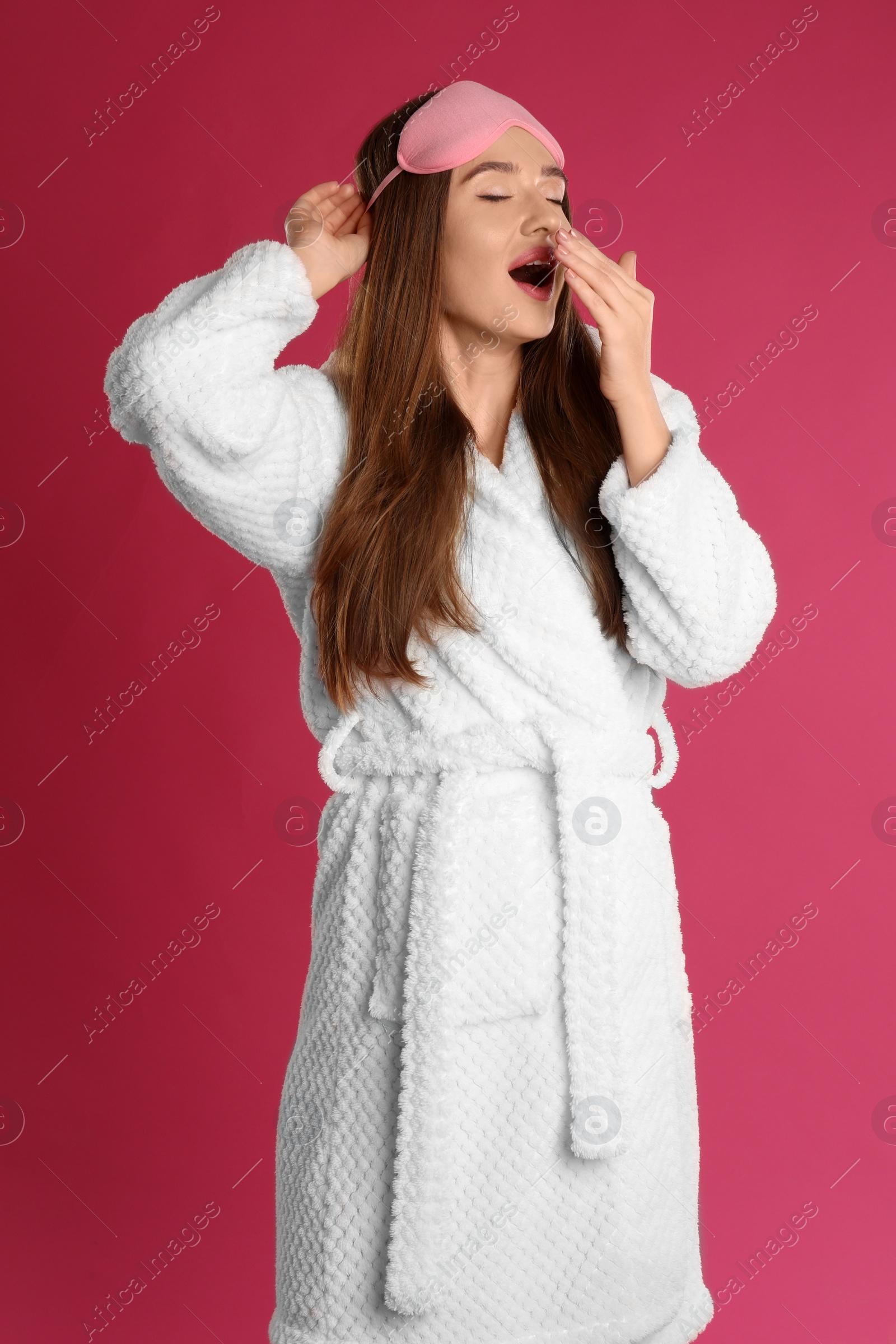 Photo of Beautiful young woman in bathrobe and eye sleeping mask yawning on crimson background