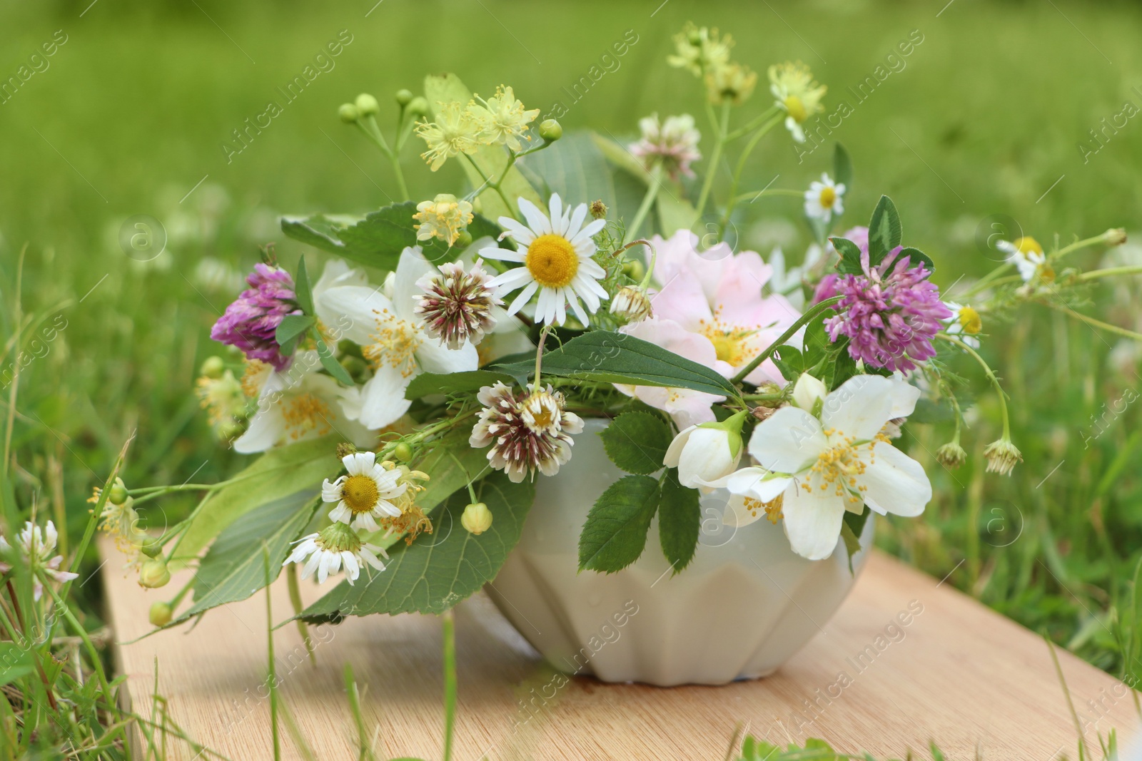 Photo of Ceramic mortar with different wildflowers and herbs on wooden board in meadow