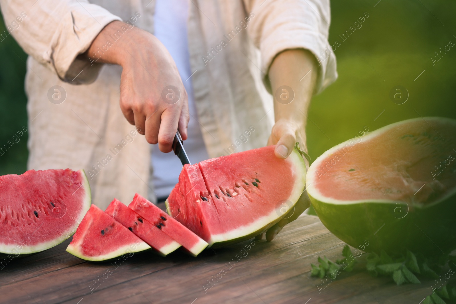 Photo of Man cutting tasty ripe watermelon at wooden table outdoors, closeup