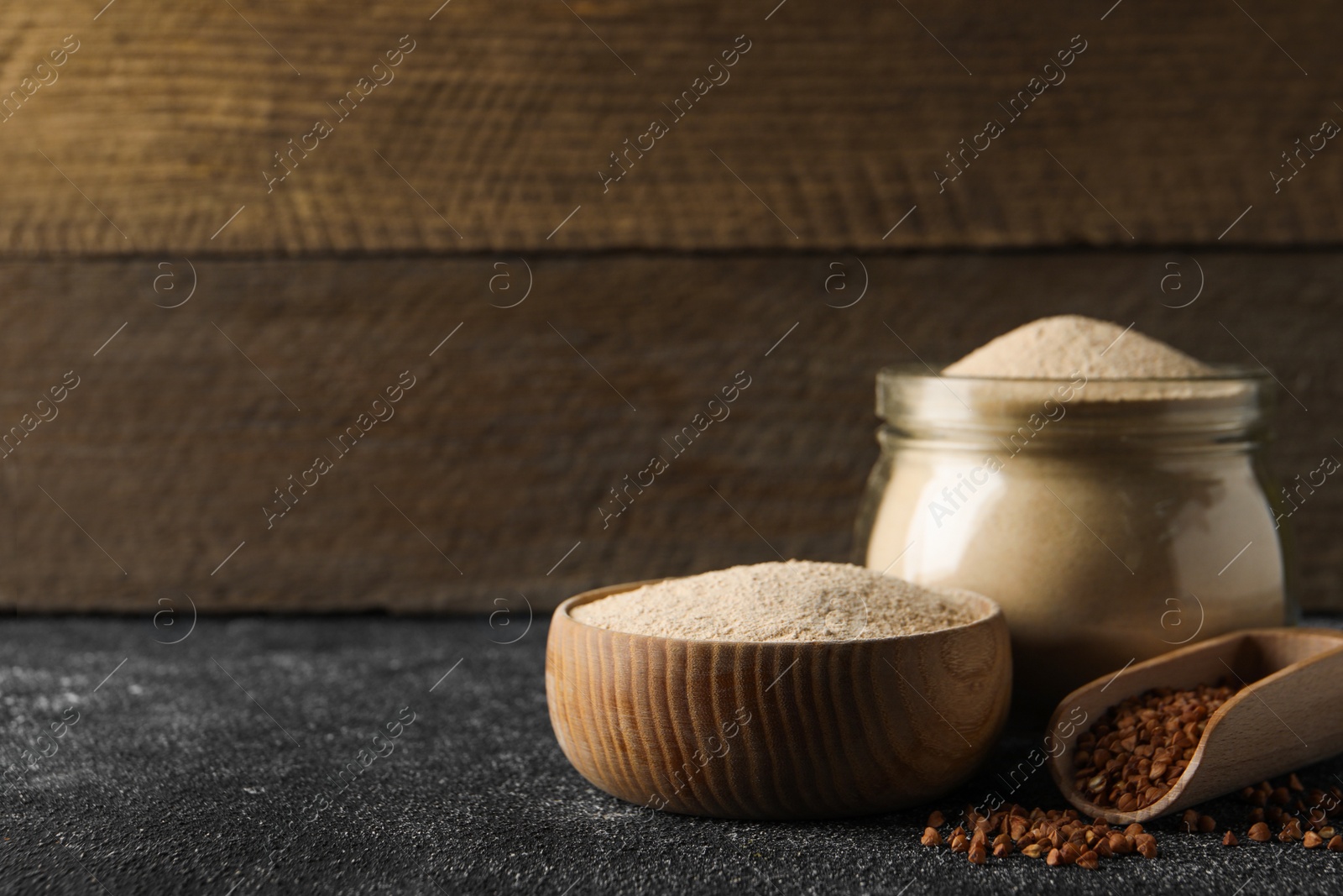 Photo of Jar and bowl of buckwheat flour near scoop with grains on black table against wooden wall. Space for text