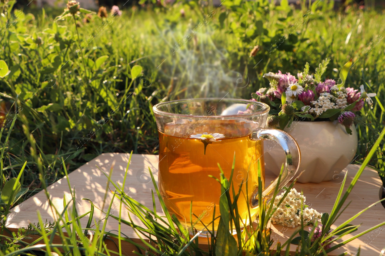 Photo of Cup of aromatic herbal tea and ceramic mortar with different wildflowers on wooden board in meadow