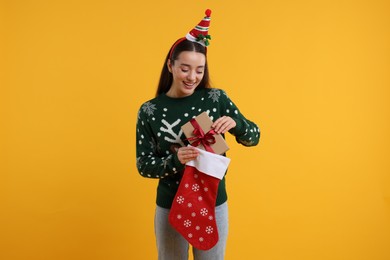 Photo of Happy young woman in Christmas sweater taking gift from stocking on orange background