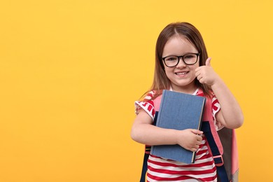 Photo of Cute little girl with backpack and book showing thumbs up against orange background. Space for text