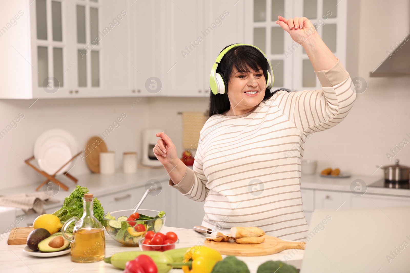 Photo of Happy overweight woman with headphones dancing while cooking in kitchen