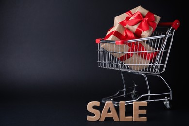 Cart with gift boxes and word Sale made of wooden letters on dark background, space for text. Black Friday