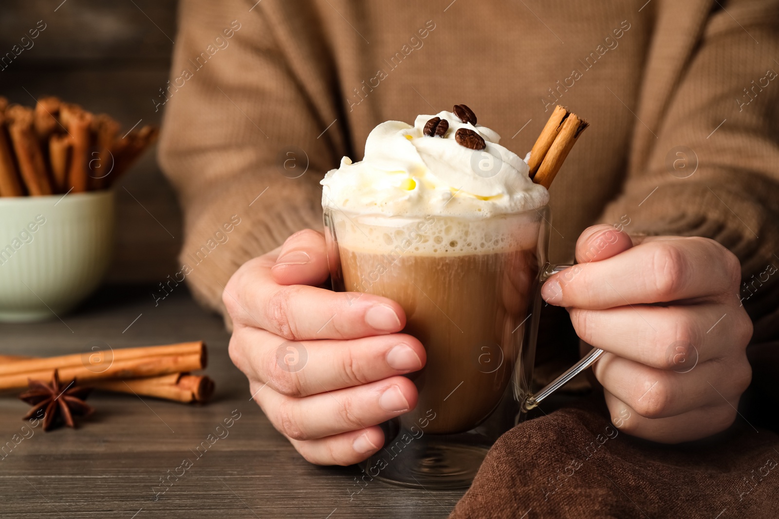 Photo of Woman holding cup of delicious coffee with whipped cream and cinnamon at wooden table, closeup